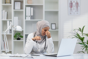 A Muslim female doctor in a hijab sitting at her office desk, feeling stressed and showing signs of...