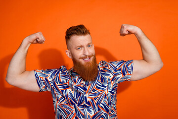 Photo of cool cheerful guy dressed print shirt rising fists showing muscles isolated orange color background