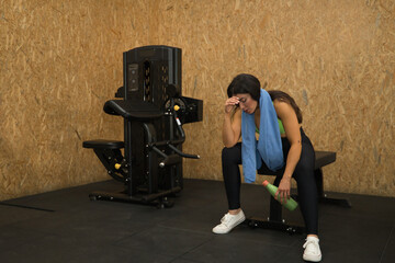 Fototapeta na wymiar Young, beautiful, brunette woman with yellow top, black leggings and towel around her neck, tired, exhausted, depressed and frustrated, sitting on a bench in the gym. Fitness concept, gymnastics.
