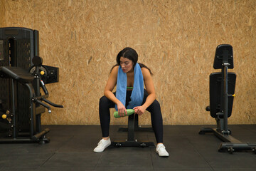 Young, beautiful, brunette woman with yellow top, black leggings and towel around her neck, tired and exhausted after exercise, sitting on a bench in the gym. Fitness concept, gymnastics, bodybuilding