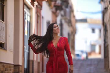 Young, beautiful brunette woman in an elegant red dress, walking while touching her hair, at dawn down a lonely street of a beautiful white Andalusian village. Concept beauty, fashion, trend, travel.