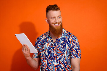 Photo of good mood guy with red beard wear print shirt hold tablet look at promo empty space isolated on vivid orange color background