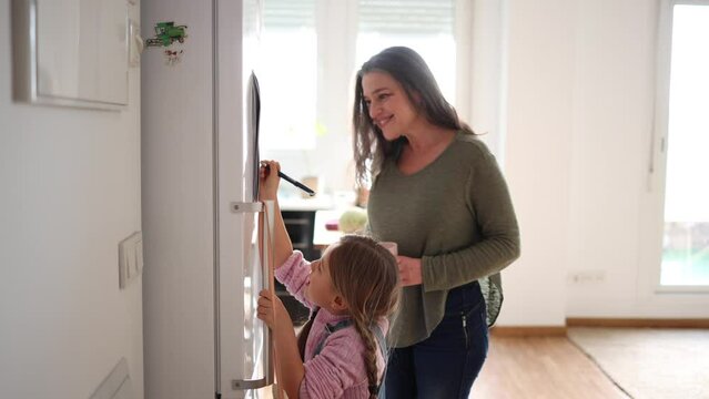 Little girl writing on a calendar on the fridge with her smiling grandmother at home