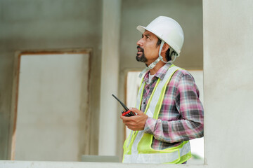 Asian builder with white helmet looks on, walkie-talkie in hand, reflective vest, against...