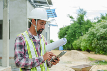 Asian Construction worker in plaid shirt and reflective vest holds blueprints and tablet, standing...