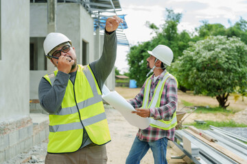 One adult male in safety vest talks on phone, making a point, another with hard hat and blueprint...
