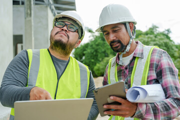 Two males in construction attire focus on a laptop, one holding tablet, with construction materials and greenery in background. Architects and engineers supervise house construction.