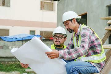 Two males in construction safety attire review a large blueprint on a building site, engaged in...