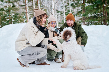 Caucasian man, woman and their daughter spending winter day in forest park sitting on hunkers and having fun with their dog