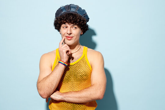 Young happy gay Latin man wear mesh tank top hat clothes put hand prop up on chin, lost in thought and conjectures isolated on plain blue cyan background studio Pride day June month love LGBT concept