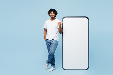 Full body young Indian man wears white t-shirt casual clothes point thumb finger on big huge blank screen mobile cell phone smartphone with area isolated on plain blue background. Lifestyle concept.