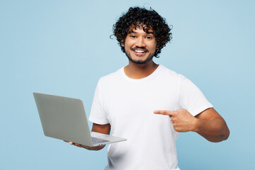 Young happy IT Indian man he wear white t-shirt casual clothes hold use work point finger on laptop pc computer isolated on plain pastel light blue cyan background studio portrait. Lifestyle concept.