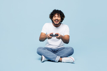 Full body young happy Indian man wear white t-shirt casual clothes sits hold in hand play pc game with joystick console isolated on plain pastel light blue cyan background studio. Lifestyle concept.