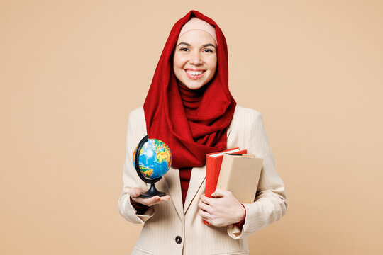 Young smart smiling happy Arabian Asian Muslim woman wear red abaya hijab suit clothes hold in hand books, globe isolated on plain beige background studio. UAE middle eastern Islam religious concept.