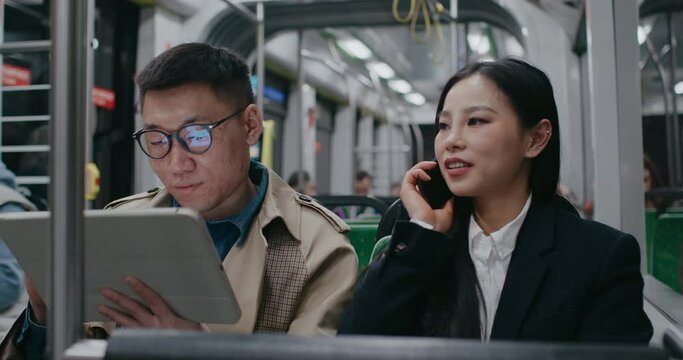 Asian man with glasses and raincoat focused in his tablet device. Young girl smiling while talking with someone. Beginning to look around. Worried about missing correct train stop. People on way home.