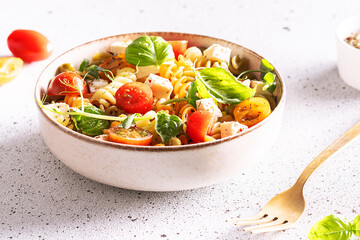 Colorful full grain fusilli pasta warm salad with feta cheese, cherry tomatoes, herbs, green pea and basil leaves on white stone background close up. Healthy diet food concept.
