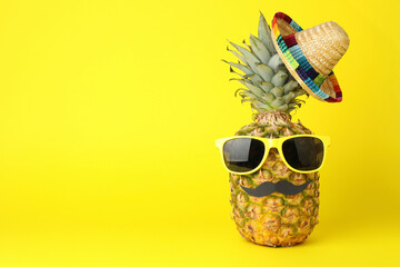 Pineapple with Mexican sombrero hat, sunglasses and fake mustache on yellow background, space for text