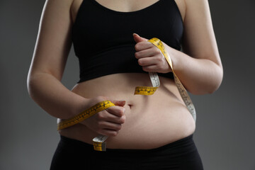 Woman measuring belly with tape on grey background, closeup. Overweight problem