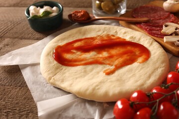 Pizza dough with tomato sauce and products on wooden table, closeup