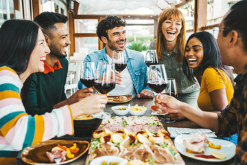 Fototapeta premium Happy friends drinking red wine sitting at restaurant table - Multiracial young people enjoying rooftop dinner party together - Food and beverage concept with guys and girls having lunch break outside