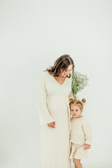 Beautiful young pregnant woman with her little 3 yearold daughter on white background. Stylish pregnant woman in beige dress. Motherhood. Mother's Day.