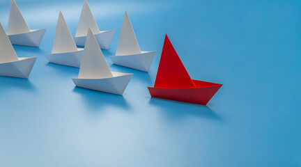 Red paper boat floating at the head of a group of white paper boats on a blue background....