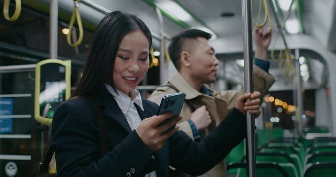 Chinese young girl standing in train or long bus. Using smartphone in public transport. Laughing from video or text. Typing something on sensor with one finger. Holding balance with left hand.