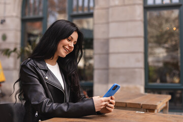 Fototapeta premium Young beautiful woman holding mobile and looking at smartphone while sitting at cafeteria. Happy girl using mobile phone, read news, shopping and look flirting. Woman smiling, sitting in cafe outdoor.