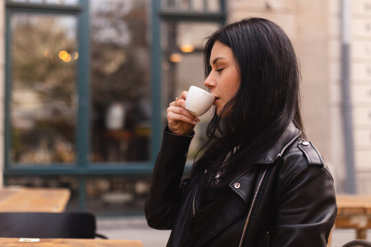 Thoughtful brunette teen woman drinking coffee in cafe outdoor, lifestyle, coffee, communication and local business, unaltered. Sensual woman sitting side. Girl wear leather jacker and look pretty.
