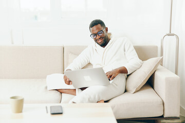 African American Man in Bathrobe Working on Laptop at Home, Smiling in the Morning This modern,...