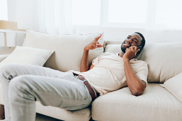 Happy African American man sitting on a black sofa, confidently using his smartphone for a video call He is dressed in a white tshirt and is comfortably relaxing at home The modern apartment serves as - obrazy, fototapety, plakaty