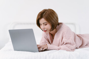 Beautiful young Asian woman in warm knitted pink clothes lying on bed while using laptop computer in bedroom. Lifestyle, comfort and technology concept. Fashion, Autumn, winter