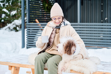 Happy Caucasian girl holding mug with hot drink sitting on bench outdoors with her lagotto romagnolo dog on winter day, copy space