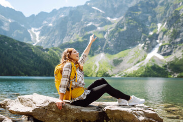 Happy tourist woman  enjoys the view of the mountain lake in sunny weather. Scenery of the majestic mountains. Active lifestyle.