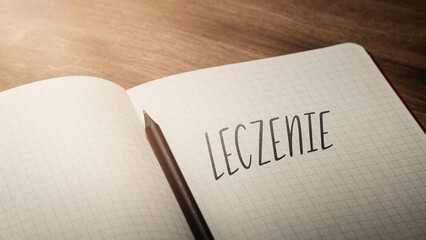  A handwritten inscription "Leczenie" on a grille of an open notebook on a wooden countertop, next to a black pencil, lighting of light. (selective focus), translation: Treatment