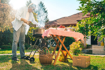 Senior adult making barbecue in backyard on a sunny day. Older man preparing a lunch outdoors while...