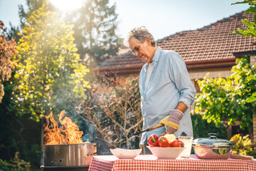 Senior adult male preparing barbecue in backyard on a sunny day. Retired man wearing shirt and...