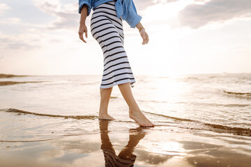 Slim female legs and feet walking along sea water waves on sandy beach. Pretty woman walks at...