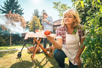 Senior blonde woman with glasses holding freshly picked tomato in her hand and looking at it, while...