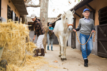 Women horsebreeders working in ranch. Senior woman leading white horse, Asian woman stacking hay.