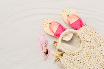 Straw bag with accessories on sand background, top view