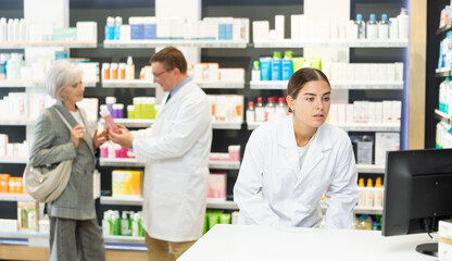 Attentive young female pharmacist looking at display of computer in chemist's shop with large...