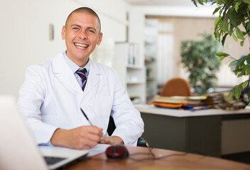 Positive middle aged man doctor therapist sitting in office at workplace and working at laptop