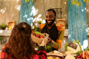 Smiling african american seller handing over variety of organic seasonal produce to caucasian...