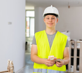 Satisfied young man in vest and helmet holding bunch of cash he obtained for first part-time job in...