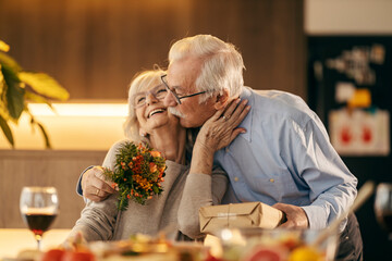 A senior man is kissing and surprising his wife with bouquet and gift on valentines day.