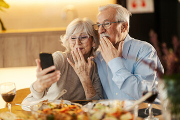 A senior couple is having video call on the phone and sending kisses while sitting at lunch table.
