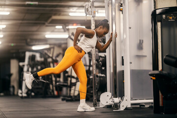 A strong black sportswoman practicing kicks on a dual pulley machine in a gym.