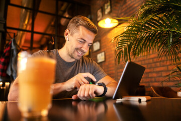 Smiling man sitting at cafe and typing on smart watch while expecting video call on tablet.