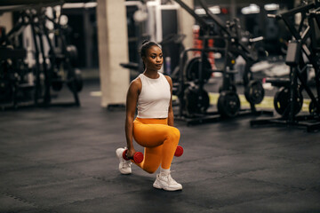 A fit black sportswoman doing lunges with dumbbells at the gym.